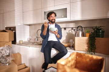 A man sits on a kitchen counter, eating from a bowl in a newly moved-in home. Surrounding him are...