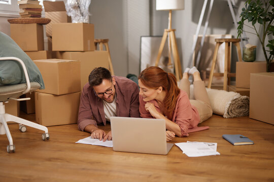 Two individuals are lying on the floor surrounded by cardboard boxes, using a laptop to strategize their moving plans and review important papers in a warm atmosphere.
