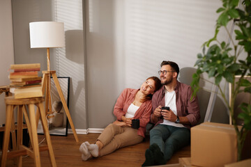 New homeowners take a moment to relax on the floor, surrounded by moving boxes and unpacked items,...
