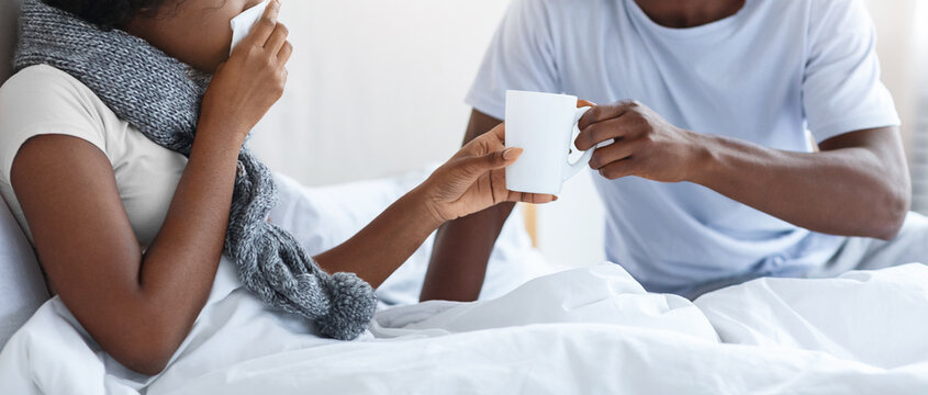 Black husband in protective face mask taking care of his sick wife, giving hot drink, bedroom interior, empty space. African american woman with scarf around neck having coronavirus, sneezing her noze