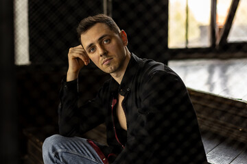 Frontal portrait of young self confident handsome young man with naked torso in a black shirt in a stylish loft interior, grey walls
