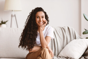 Potrait of happy millennial long-haired brunette woman in cozy outfit sitting on sofa at home,...
