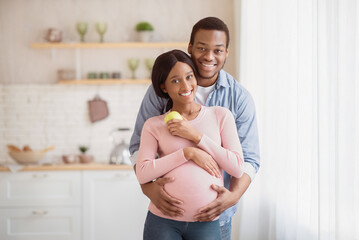Portrait of loving black husband hugging his pregnant wife at home. Beautiful expectant woman with...