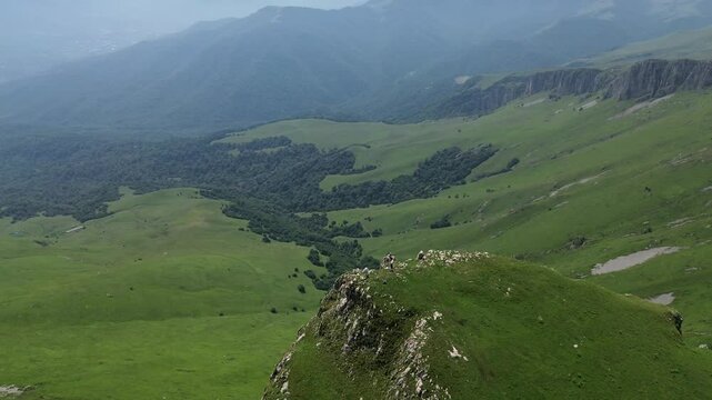 Drone panorama circling a beautiful rocky formation in mountains covered with lush green grass, set against a clear blue sky.