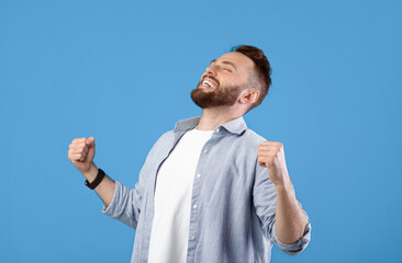 Triumphant young man gesturing YES with closed eyes on blue studio background, panorama. Bearded...