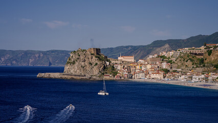 Picturesque coastal view of Scilla, Calabria, featuring Ruffo Castle, colorful buildings, and a sailboat navigating the Tyrrhenian Sea on a sunny day