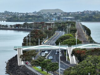 People walk and cars drive along a coastal road of Tamaki Drive and railway line with a pedestrian bridge in Auckland, New Zealand