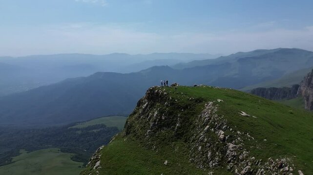 Circular drone flight around a rocky hill in green mountains with three people standing on top. Beautiful landscape and terrain set against a clear blue sky.
