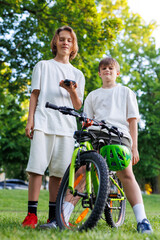 Two boys in white T-shirts pose confidently outdoors with a green mountain bike. One holds a phone, the other rests on the handlebar, both looking down with cocky expressions in summer park setting