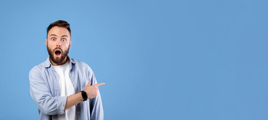 Shocked young guy standing with open mouth, pointing at empty space in disbelief, blue studio...