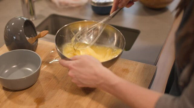 High angle detail shot of a woman hands beating eggs with a manual whisk on a grey countertop and wooden board in a home kitchen. Calm, everyday cooking task. Horizontal, 4K
