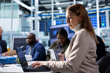 Factory employee works on laptop next to executives discussing solar panel automation strategies. Worker typing on notebook next to management team enhancing sustainable manufacturing processes