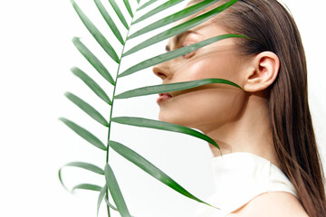Stylish young woman with long hair partially hidden behind a palm leaf, showcasing natural beauty and elegance in a minimalist setting.
