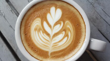 A close-up of a latte in a white cup, featuring intricate latte art resembling a leaf. The cup is placed on a rustic wooden table. - Powered by Adobe