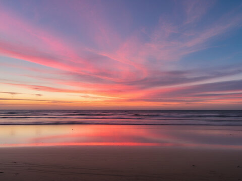 A serene landscape photo capturing a vibrant beach sunset with a sky full of pink and purple clouds reflecting on the wet sand and calm ocean.