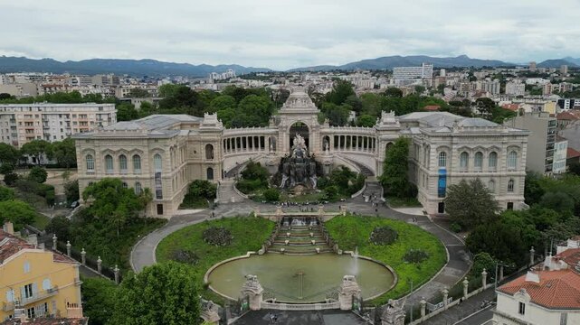 Aerial view of Palais Longchamp in Marseille, France. The grand architecture features a central fountain surrounded by lush greenery and pathways. The city skyline is visible in the background.