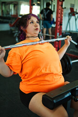 Beautiful, overweight Latina woman concentrating on lifting weights on an exercise machine at a gym. Wearing colorful clothes, hair, and retro makeup, she's positive. She's pushing herself.