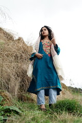 A female Kashmiri model wearing a traditional pheran and shawl, posing for the artisanal brand Kaar Kash, with a richly embroidered curtain as a backdrop in a serene outdoor setting.

