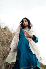 A female Kashmiri model wearing a traditional pheran and shawl, posing for the artisanal brand Kaar Kash, with a richly embroidered curtain as a backdrop in a serene outdoor setting.


