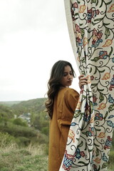 A female Kashmiri model wearing a traditional pheran and shawl, posing for the artisanal brand Kaar Kash, with a richly embroidered curtain as a backdrop in a serene outdoor setting.

