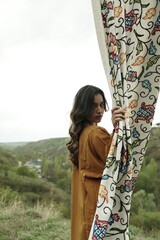 A female Kashmiri model wearing a traditional pheran and shawl, posing for the artisanal brand Kaar Kash, with a richly embroidered curtain as a backdrop in a serene outdoor setting.

