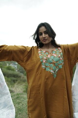 A female Kashmiri model wearing a traditional pheran and shawl, posing for the artisanal brand Kaar Kash, with a richly embroidered curtain as a backdrop in a serene outdoor setting.

