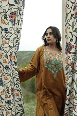 A female Kashmiri model wearing a traditional pheran and shawl, posing for the artisanal brand Kaar Kash, with a richly embroidered curtain as a backdrop in a serene outdoor setting.

