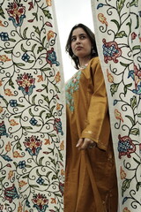 A female Kashmiri model wearing a traditional pheran and shawl, posing for the artisanal brand Kaar Kash, with a richly embroidered curtain as a backdrop in a serene outdoor setting.

