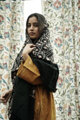 A female Kashmiri model wearing a traditional pheran and shawl, posing for the artisanal brand Kaar Kash, with a richly embroidered curtain as a backdrop in a serene outdoor setting.

