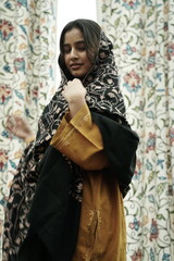 A female Kashmiri model wearing a traditional pheran and shawl, posing for the artisanal brand Kaar Kash, with a richly embroidered curtain as a backdrop in a serene outdoor setting.

