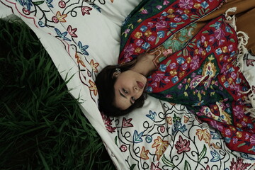 Woman lying peacefully on a traditional carpet spread over lush green grass, blending comfort,...