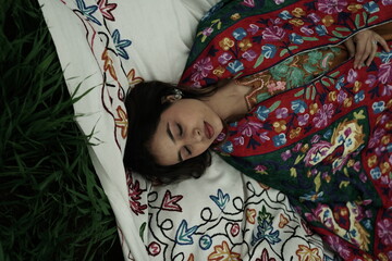 Woman lying peacefully on a traditional carpet spread over lush green grass, blending comfort,...