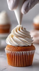 A golden cupcake is being topped with buttercream by gloved hands, the swirl halfway done. Natural light highlights its texture on a clean white surface. 
