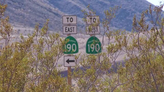 Cars slow down and stop by California Route 190 signs framed by desert creosote bushes in Death Valley National Park.