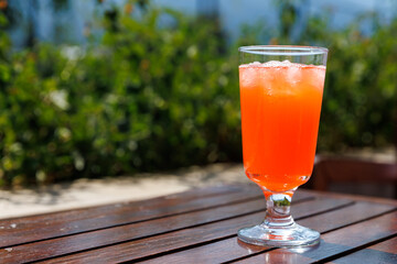 Bright orange drink served in glass with ice cubes on wooden table outdoor with blurred green foliage in background, summer concept, copy space.