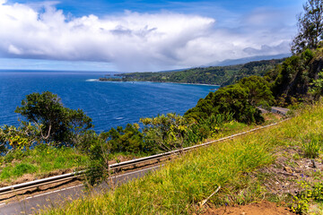 Scenic coastal road view with lush greenery, blue ocean, and cloudy sky.