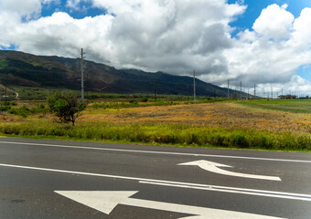 Road with arrows pointing in opposite directions, field, mountains, and cloudy sky.