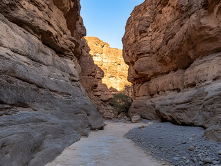 Narrow canyon with rocky walls and sunlit cliffs under clear blue sky, dry desert landscape with sandy and gravel path, natural heat entrapment creating warm atmosphere