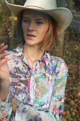 Attractive young woman in cowboy hat with invisible cigar or cigaret in hand, while looking away from camera. Natural no make up beauty in autumn environment. Smoking concept
