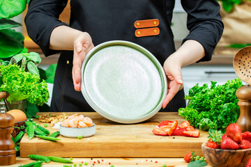 Chef cooking food in the kitchen. Cook holds ceramic plate in hands for prepare salad., wooden table with kitchenware, vegetables, herbs and ingredients. Healthy vega eating, recipes, blogging
