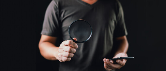 A man holds a magnifying glass and a smartphone against a black background, and suggests detailed...