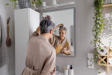An elderly woman in her ages, a pensioner in silk pajamas, examines her gray hair holding a comb.