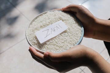Zakat Rice or zakat fitrah. Rice grains in a glass bowl with zakat written on paper. Islamic concept of zakat to help the poor in need. held by a woman's hand