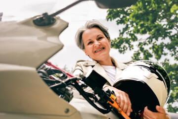 Smiling senior woman with gray hair holding helmet next to motorcycle, confident and free outdoors