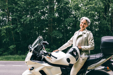 Stylish senior woman with gray hair sitting on motorcycle holding helmet near forest road