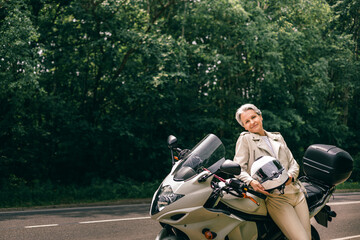Stylish senior woman with gray hair and motorcycle posing on roadside, symbol of freedom and grace