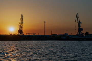 Obraz premium Port cranes for cargo transshipment on the mooring line of the Astrakhan port on the right bank of the Volga River against the background of the setting sunб Astrakhan, Russia