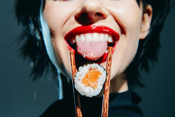 Young woman enjoying sushi with chopsticks, vibrant red lips and playful expression, against a dark modern background.