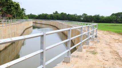 Metal fence beside irrigation canal. Close up of safety fence installed on cement edge of irrigation canal in rural Thailand on blue sky and green rice field background with copy space. selective focu
