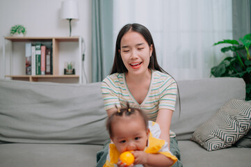 Portrait of Asian mother sits on the sofa holding her little daughter and playing with her happily in the living room at home.